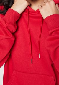 Osoba poprawiająca sznurki kaptura jasnoczerwonego hoodie z żebrowanymi mankietami i przednią kieszenią.