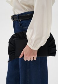 Černá kabelka typu clutch s nařaseným saténovým detailem, držena v ruce. Má obdélníkový tvar a hladkou texturu látky. Neformální outfit s džíny.