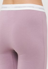 Lyse lilla leggings lavet af blødt stof, med en hvid talje med teksten "COPENHAGEN STUDIOS." Glat tekstur med tætsiddende design.