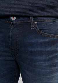 Primer plano de unos pantalones vaqueros de mezclilla azul oscuro con costuras naranjas y una bragueta de botones, combinados con un suéter de canalé oscuro.