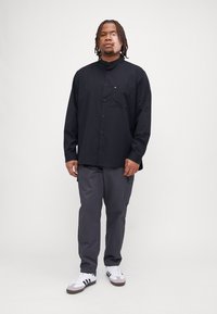 Camicia nera con bottoni, colletto e tasca, abbinata a pantaloni cargo grigi e scarpe bianche con strisce nere. Look casual, vestibilità rilassata.