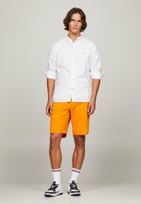 Hvid skjorte med knapper, optrukne ærmer, parret med lysorange shorts. Sort-hvide sneakers med stribede ankelstrømper. Neutral baggrund.