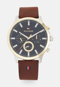 Orologio con cinturino in pelle marrone, lunetta dorata, quadrante blu navy, dettagli in argento e tre sottocronografi. Presenta il logo del marchio e cuciture rosse, bianche e blu.