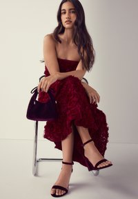 Rochie fără bretele burgundă cu textură florală, asortată cu o mini geantă de catifea și pantofi negri cu bretea la gleznă, așezată pe un scaun cromat.