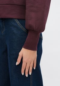 Mørkerød genser med puffede ermer, ribbestrikkede mansjetter og rundet kant, kombinert med mørkeblå jeans med gule sømmer.