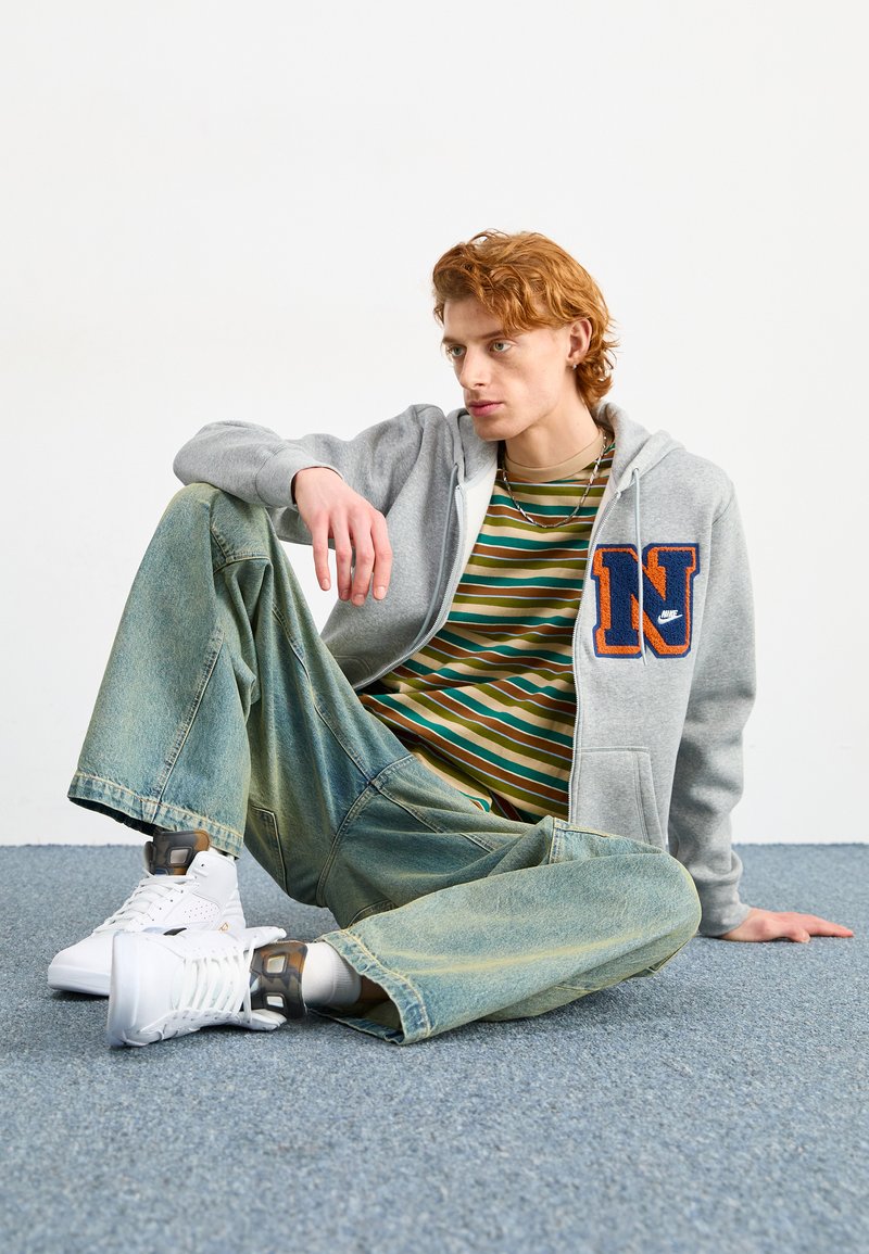 Jasnoszara bluza z kapturem z logo "N", zielona i żółta koszulka w paski, oversize'owe jasnoniebieskie dżinsy oraz białe sneakersy z szarymi akcentami.