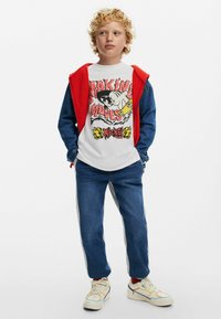 White T-shirt with red graphic design, red zip-up hoodie, denim jacket, blue jeans with gray side stripes, and colorful sneakers.