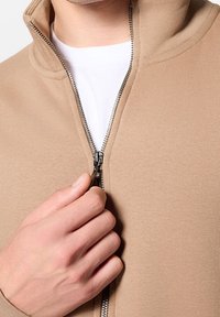 Felpa con zip beige in tessuto texture, dotata di zip argentata, colletto alto e una maglietta bianca visibile sotto.