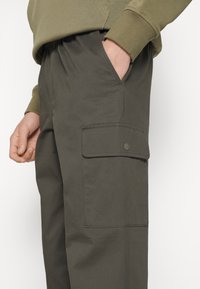 Cargo byxor i mörk olivgrön färg, med resårmidja, sidoficka och en lapplås ficka med knappstängning. Slätt, lätt material.