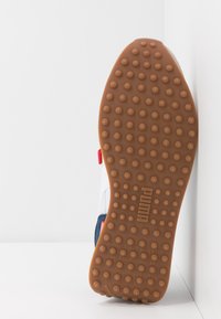Boty Puma s hnědou gumovou podešví se vzorem kruhových vroubků, bílým svrškem, námořnicky modrými akcenty a červeným detailem na patě.