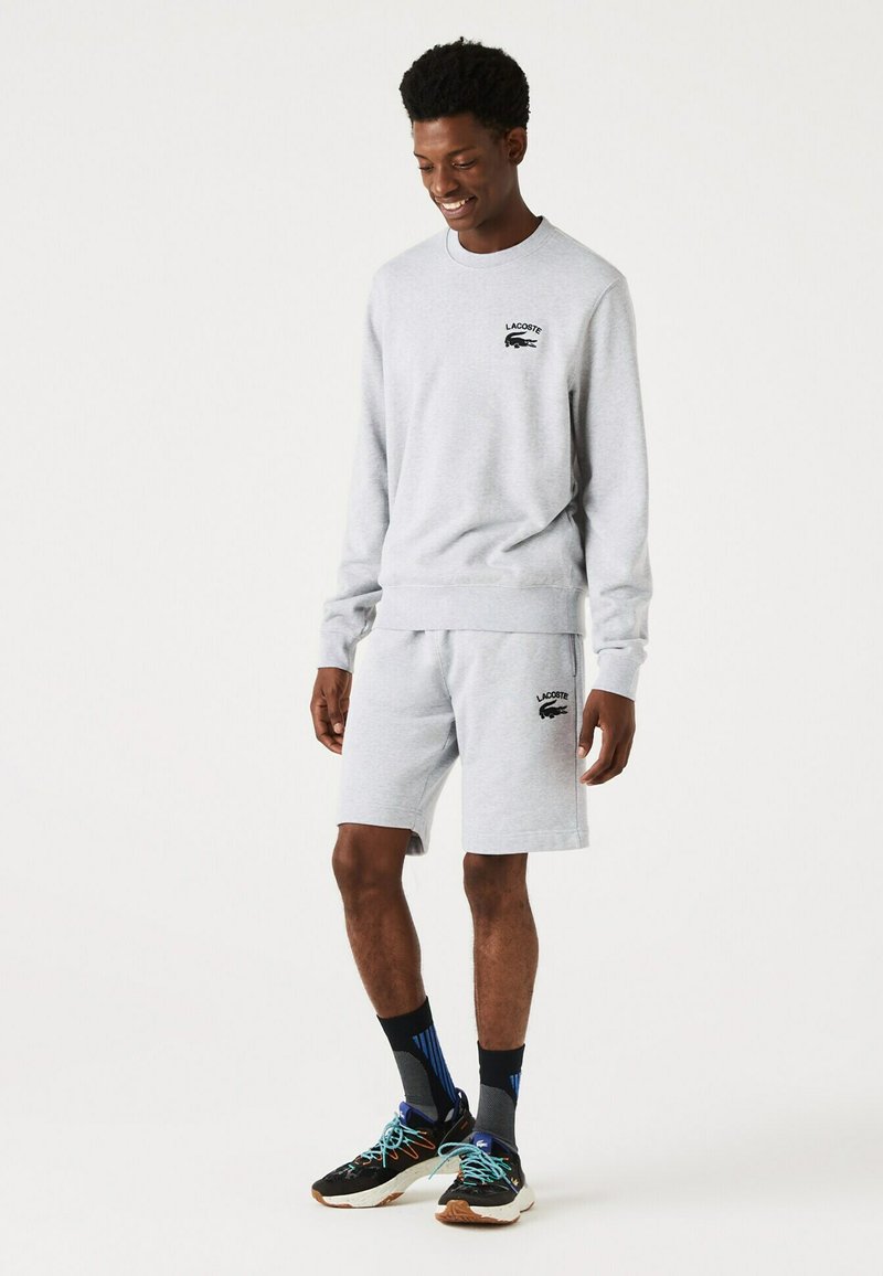 Homme portant un sweat à capuche Lacoste gris clair et un short assorti, debout et souriant, avec des chaussures de sport noires et bleues et des chaussettes.