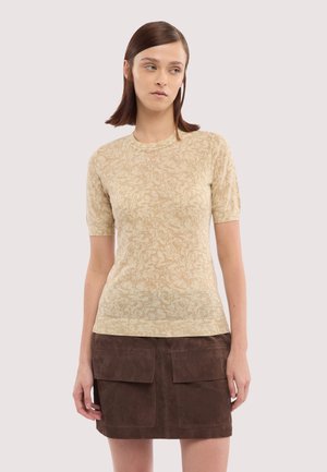 Jeune femme portant un haut beige à manches courtes avec un motif et une jupe en daim marron avec des poches avant, debout devant un fond uni.