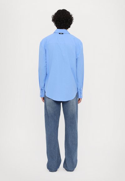 Hellblaues Langarmhemd mit Kragen, aus glattem Stoff, mit Rückennähte und abgerundetem Saum, kombiniert mit weiter geschnittenen hellblauen Jeans.