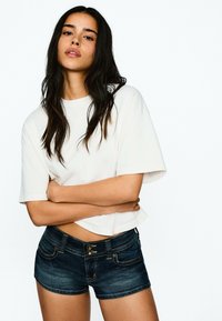 White cropped t-shirt, short sleeves, paired with dark denim shorts featuring two button closures and contrasting stitching on pockets.