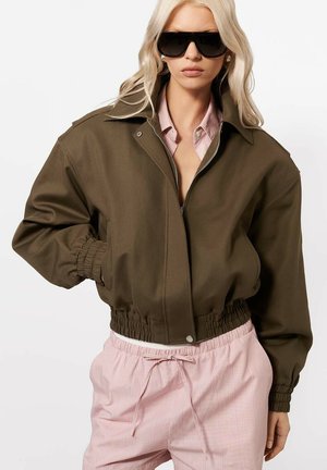 Kobieta w za dużej oliwkowozielonej kurtce, różowych spodniach w paski i dużych czarnych okularach przeciwsłonecznych, pozująca z jedną ręką w kieszeni.