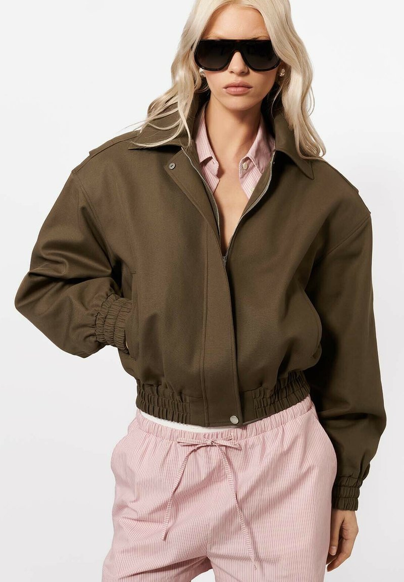 Femeie purtând o jachetă supradimensionată verde măsliniu, pantaloni roz cu dungi și ochelari de soare negri mari, pozând cu o mână în buzunar.