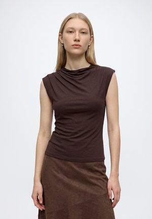 Femme aux cheveux blonds raides portant un haut marron foncé sans manches et une jupe marron assortie, debout devant un fond blanc uni.