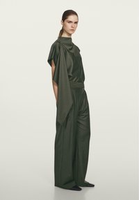 Kobieta z włosami związanymi z tyłu, ubrana w ciemnozielone, oversize'owe spodnie z szerokimi nogawkami oraz pasującą asymetryczną bluzkę z opadającym rękawem.