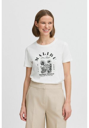 T-shirt en coton blanc avec un imprimé graphique noir portant "MALIBU", des palmiers, et le texte "TROPICAL HOLIDAY", associé à un pantalon beige.