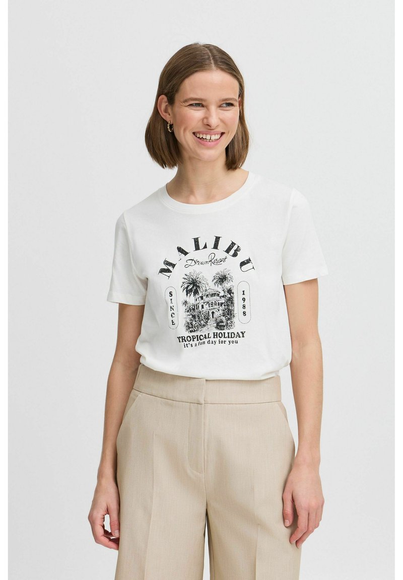 T-shirt en coton blanc avec un imprimé graphique noir portant "MALIBU", des palmiers, et le texte "TROPICAL HOLIDAY", associé à un pantalon beige.