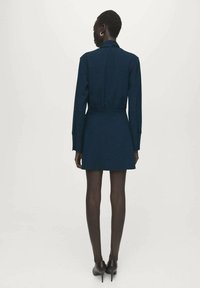 Donkerblauwe jurk met lange mouwen, een getailleerde taille en een kraag. De stof lijkt gestructureerd. Gecombineerd met zwarte pumps.