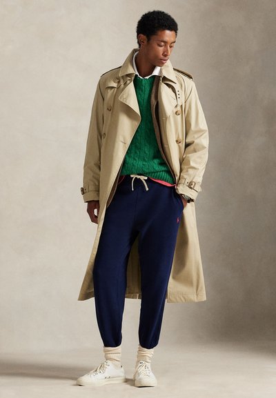 Jonge man met een beige trenchcoat, groene trui, donkerblauwe joggingbroek en witte sneakers, staand met handen in de zakken en naar beneden kijkend.