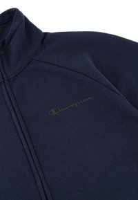 Giacca blu navy con zip, realizzata in tessuto morbido, caratterizzata da un alto colletto, cuciture evidenti e un piccolo logo di Champion in una tonalità più chiara.