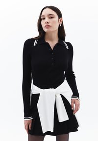 Svart ribbad långärmad polo topp med en vit randig krage, kombinerad med en svart kjol och en vit tröja knuten i midjan.