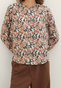 Blusa floreale con base bianca, motivi arancioni e verde acqua. Chiusura con bottoni sul davanti, maniche lunghe, scollo arrotondato e tessuto liscio.