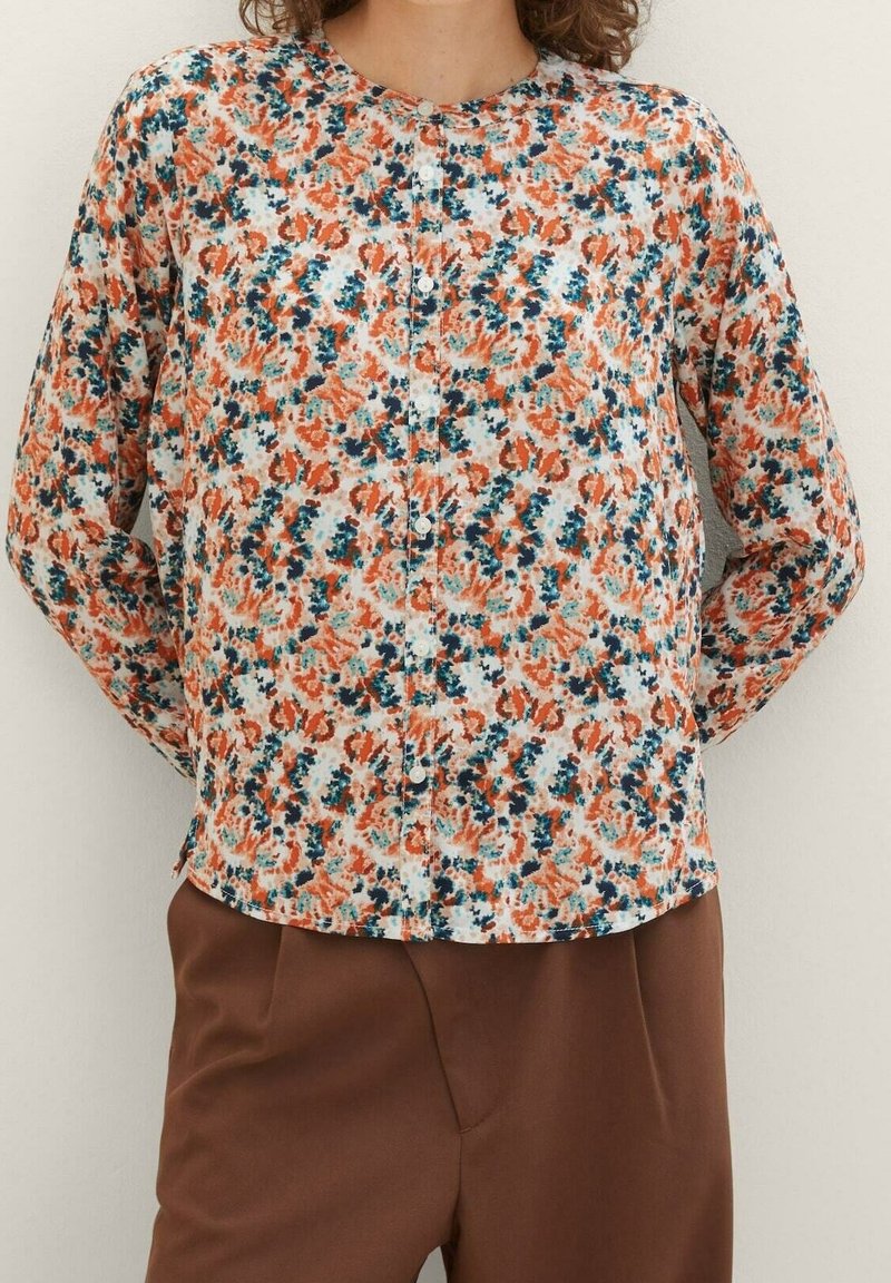 Bloemenblouse met een witte basis en patronen in oranje en groenblauw. Geknoopt aan de voorkant, met lange mouwen, een ronde halslijn en een gladde textuur.