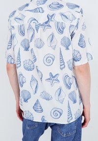 Bílá košile s krátkým rukávem s modrými vzory lastur a hvězdic. Vyrobena z lehké látky, s volným střihem a límečkem.