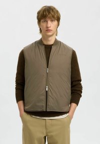 Gepolsterte Weste in gedämpftem Braun, mit einem Frontreißverschluss, geripptem Kragen und Seitentaschen, übereinander getragen mit einem dunklen Pullover.