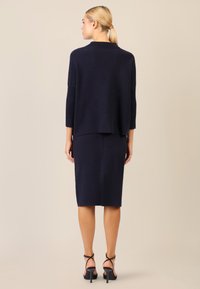 Granatowy sweter z dzianiny z szerokimi rękawami i prążkowanymi detalami, w zestawieniu z dopasowaną granatową spódnicą, podkreśloną czarnymi sandałami na obcasie.