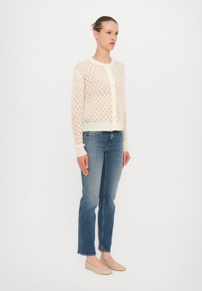 Cardigan lavorato a maglia di colore crema con un motivo testurizzato, dotato di scollatura tonda e bottoni frontali, abbinato a jeans blu e scarpe basse chiare.