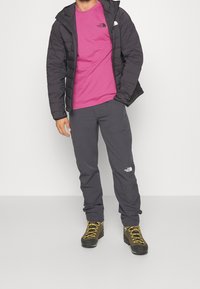 Černá zateplená bunda přes růžové tričko, šedé kalhoty a boty se žlutými tkaničkami. Obsahuje loga značek na bundě a tričku.