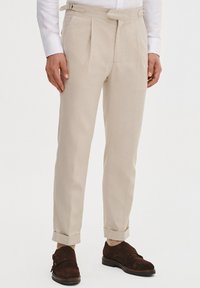Beżowe dopasowane spodnie z podwójnymi zakładkami, szlufkami na pasek i rolowanymi mankietami, wykonane z lekkiego materiału, w połączeniu z ciemnobrązowymi zamszowymi butami.