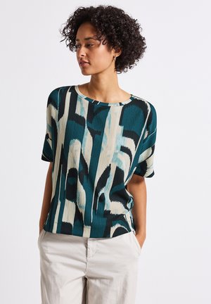T-shirt à manches courtes avec un motif abstrait bleu-vert et crème, texture douce, encolure ronde et coupe décontractée. Associé à un pantalon clair.
