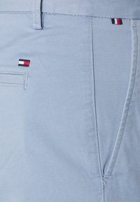 Ljusblått tyg i närbild som visar en ficka och två små rektangulära logotags i röd, vit och marinblå.