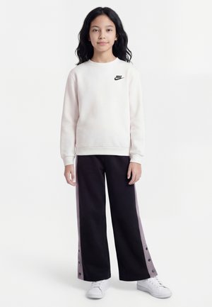 Nuori henkilö seisoo valkoisessa Nike-hupparissa, mustissa leveälahkeisissa housuissa, joissa on sivunapit, ja valkoisissa lenkkareissa valkoista taustaa vasten.