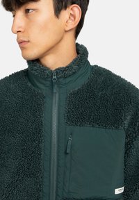 Fleecejacka i mörkgrön med hög krage, dragkedja och ett texturerat, fluffigt tyg. Har en dragkedjeförsedd bröstficka och släta accenter.