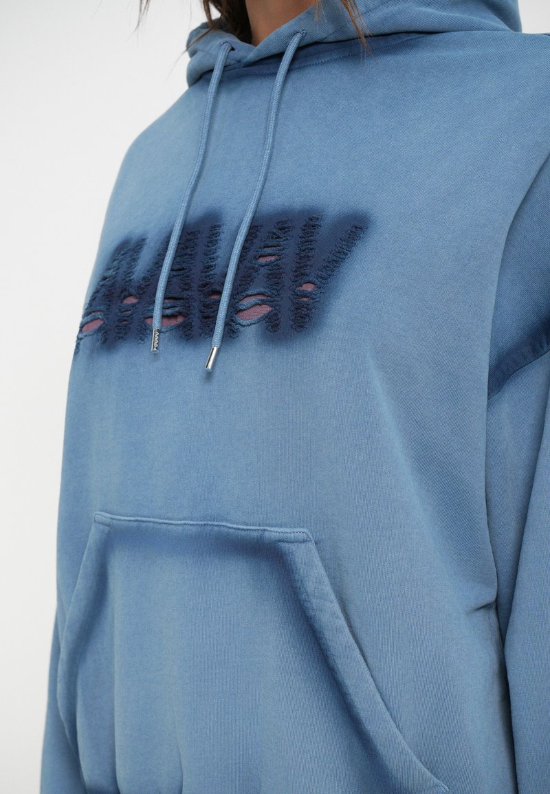 Fadede blå hættetrøje med frontlomme, snøring i hætten og slidte bogstaver i mørkeblå. Blødt bomuldsstof og afslappet pasform.