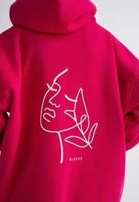 Fuchsia hoodie met een witte lijntekening van een gezichtillustratie en de tekst "MIRONS" eronder. Zacht materiaal en een relaxed fit ontwerp.