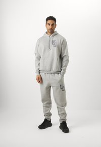 Conjunto de sudadera con capucha y pantalones joggers grises, confeccionado con tejido suave, que presenta un bolsillo en la parte frontal y detalles de logotipo. Combinado con zapatillas deportivas negras.