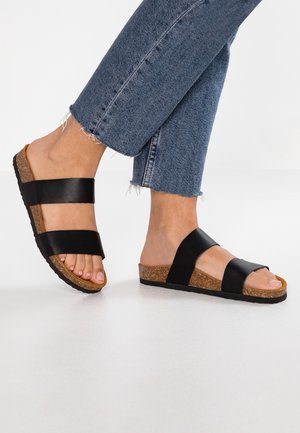 Schwarze Ledersandalen mit zwei breiten Riemen, Korkfußbett und strukturierter Gummisohle. Getragen zu hochgekrempelten blauen Jeans.