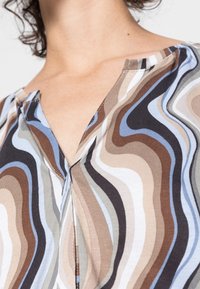Top multicolore con scollatura a V caratterizzato da motivi a onda nei colori marrone, blu, crema e nero, realizzato in un tessuto morbido. I dettagli delle cuciture sono visibili.