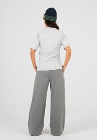 Persona che indossa un berretto blu navy, una maglietta grigia chiara a maniche corte e pantaloni grigi a gamba larga, in piedi con le mani sui fianchi e rivolto di spalle.