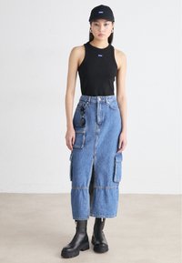 Black sleeveless top, blue denim midi skirt with cargo pockets, front slit, paired with black heeled boots and a black cap.