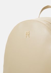 Zaino beige con una texture liscia, caratterizzato da un logo dorato sulla parte frontale e dettagli cuciti lungo i bordi. Forma rotonda e design minimale.