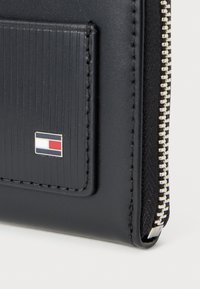 Sort læderpunge med teksturerede lodrette linjer, sølv lynlåslukning og et lille logo i rød, hvid og blå på frontlommen.