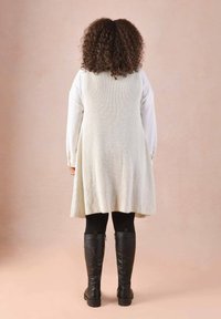 Long, light grey knit vest with a flared silhouette, worn over a white shirt and black leggings. The model wears knee-high black boots.